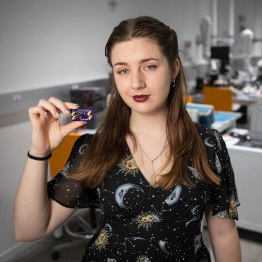 Harvard SEAS senior Molly Boswroth wearing a black shirt with moons, suns and stars, holding a computer chip in front of electrical engineering equipment