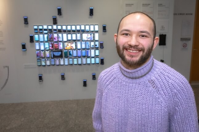 A man wearing a lavender sweater and silver chain standing next to a wall of smartphones displaying the same video