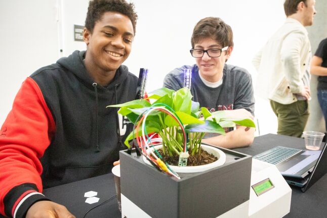 Two male Harvard students sitting behind a green houseplant connected to an automatic watering system