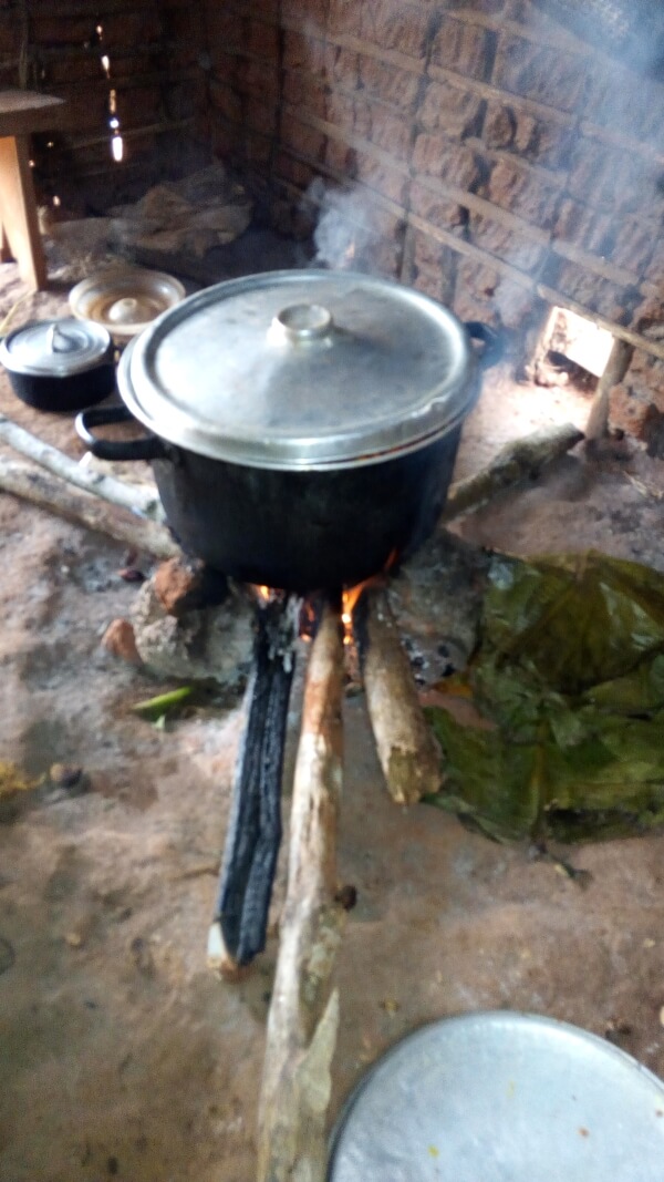 A black cooking pot with a metal lid rests on top of several pieces of firewood and rocks. There is a fire burning underneath, causing steam to emit from the pot.