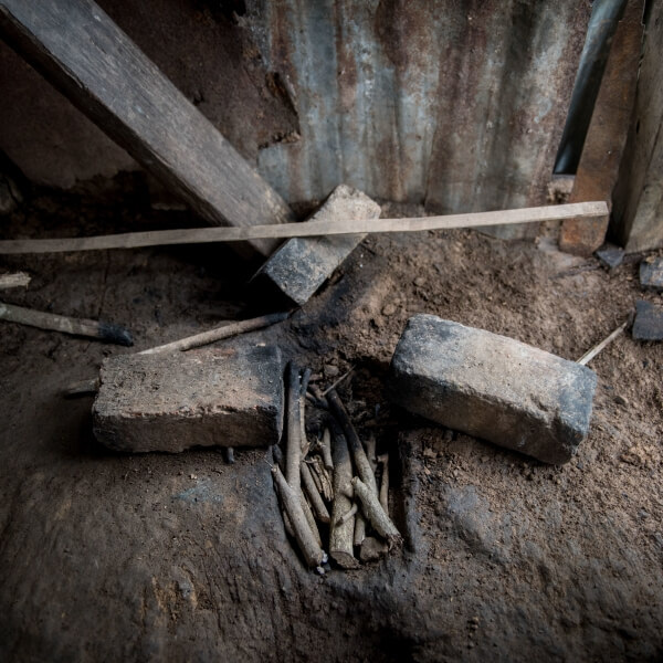 Two large bricks lay almost side-by-side on the ground (presumably for holding up cooking pots). In between them is a hole in the dirt filled with wooden sticks for burning. There is some wood and a brick scattered along a surrounding metal sheet wall.