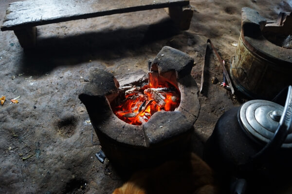 A cylindrical wooden stove burns firewood. There is a metal cooking pot in the bottom right corner. A wooden bench is nearby. 