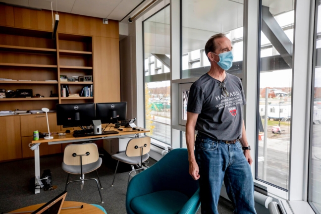 Dean Doyle looks outside the window of his new office space, which includes a built-in bookshelf and multiple tables. He is dressed casually in a gray SEAS t-shirt and jeans.