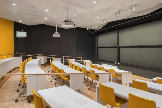 A lecture-style classroom in the SEC contains bright yellow chairs behind long tables, all facing towards a floor-to-ceiling chalkboard area.