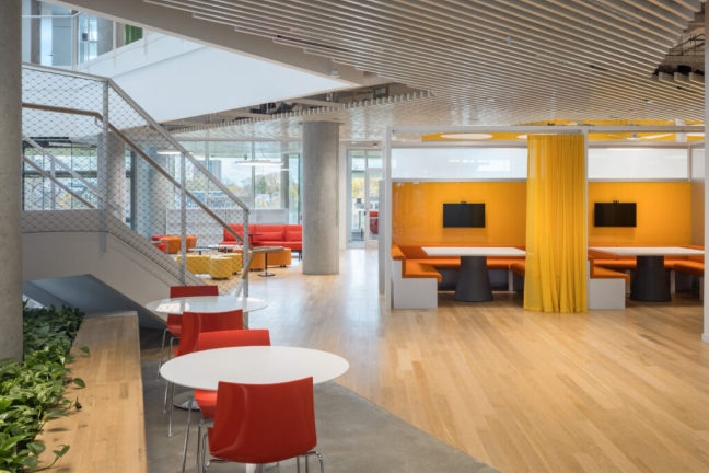 Small private workstations and round tables decorate a common area of the SEC. The chairs and benches are bright orange and yellow. Some plants are in the foreground. 