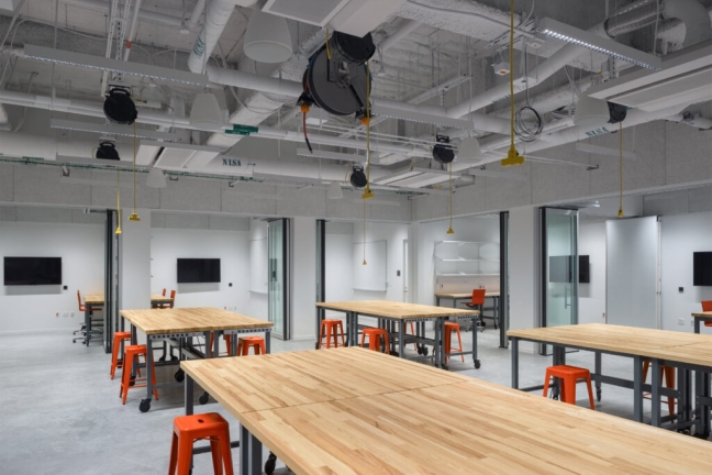 A lab in the SEC contains bright orange stools and massive wooden work tables. Ventilation tubes cover the ceiling and there are several small workstations along the walls.