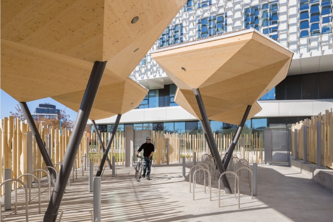 An outdoor section of the SEC contains large wooden sculptures that shade and separate a bike parking area. A man in cycling gear walks his bike towards one of the spots.
