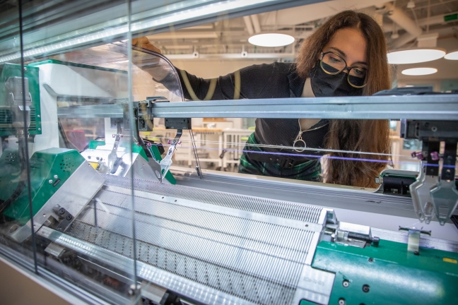 Vanessa Sanchez stands behind a large white and green machine that is creating knit material.