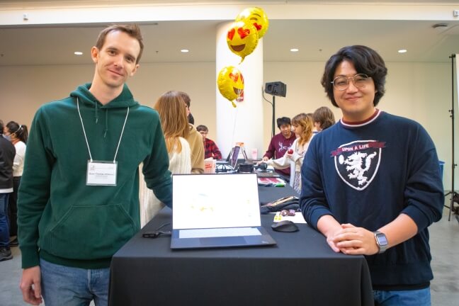 Two male Harvard students standing beside a laptop at a computer science festival in Cambridge