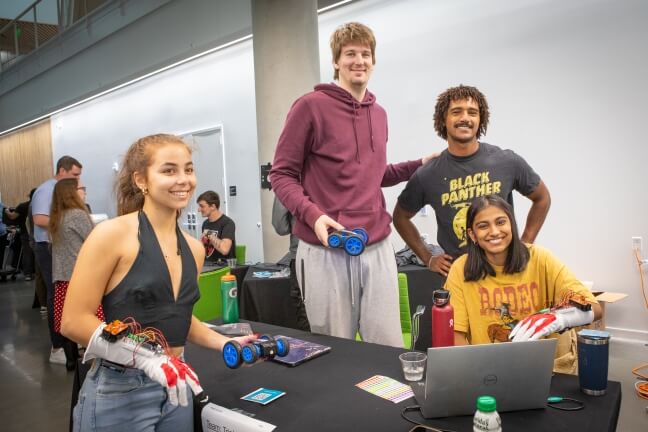 Four Harvard students holding toy cars controlled using white gloves covered in wires and electronic parts