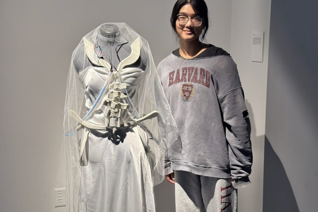 A Harvard student standing next to a wedding dress with colored LED light strips and 3D-printed spinal fragments glued onto it