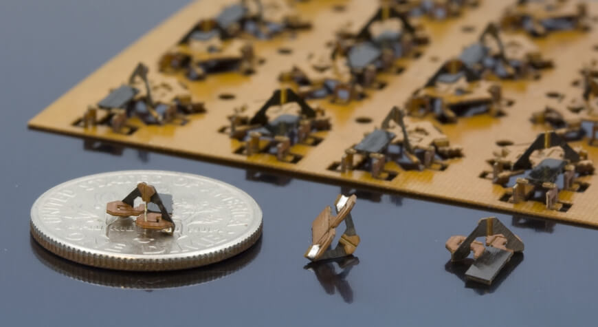 Several black and brown microrobotic mechanisms are printed on a sheet. One of the mechanisms sits on top of a coin, showcasing its miniature size.