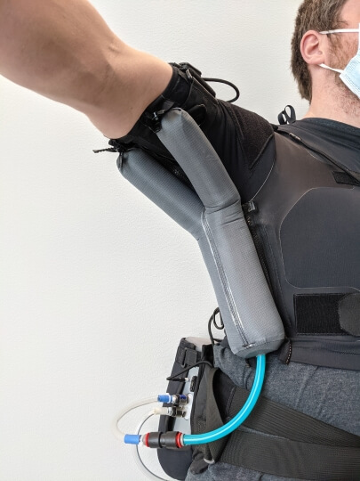 Gray balloon-like actuators cover a person's underarm in a Y-shape and inflate to help that person raise their arm.