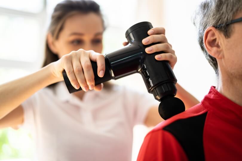 A person in a white polo holds a black massage gun to someone's right shoulder in the foreground.