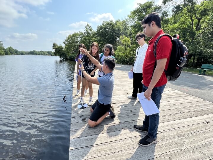Harvard SEAS assistant director Bryan Yoon kneels on a dock over the Charles River while high school students watch him