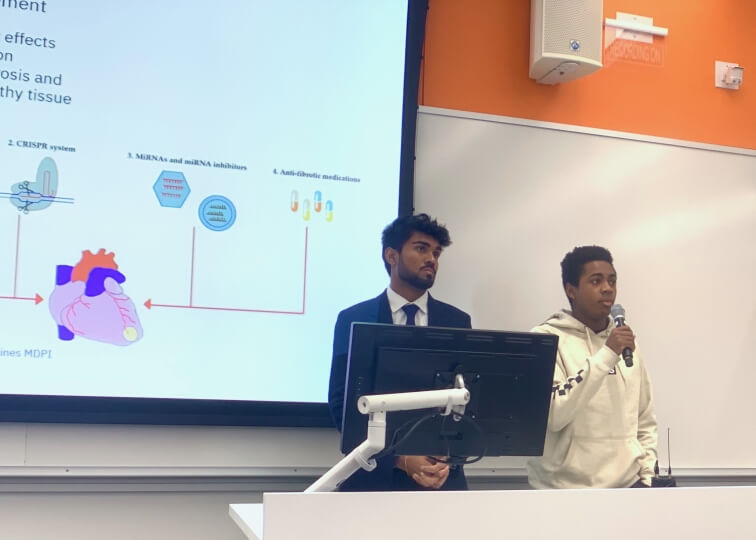 Two male Harvard students presenting behind a computer monitor, in front of a digital projection screen