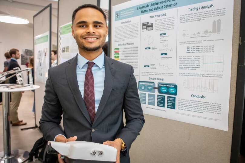 Harvard SEAS senior Adam Reid wearing a suit and tie in front of his senior capstone project poster, holding an air pollution and traffic monitor