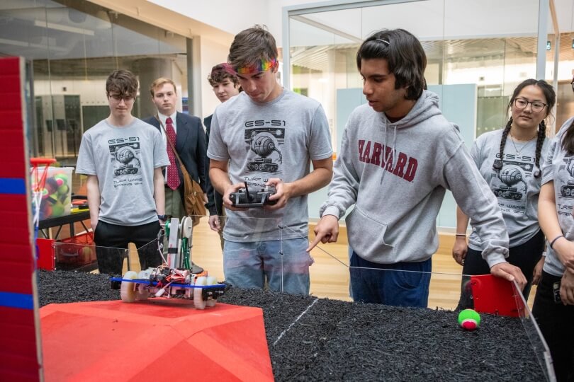 Two male Harvard students drive a small robot up a ramp in front of a group of onlookers