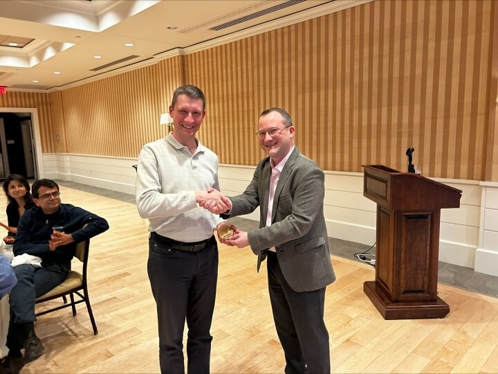 Two men smiling and shaking hands on a stage, one receiving a trophy from the other, with an audience in the background in a conference hall.