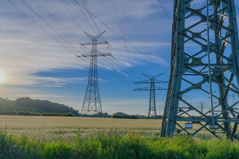 Power lines and electrical pylons stretch across a grassy field under a blue sky with scattered clouds.