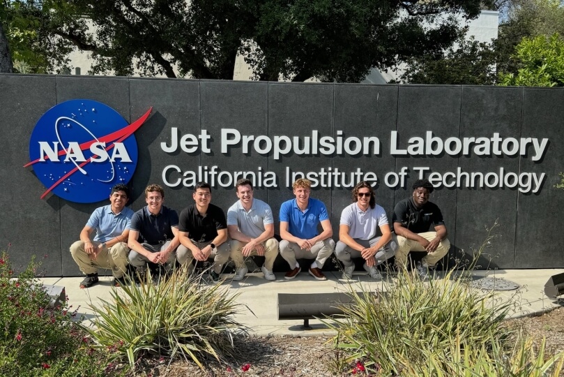 Harvard SEAS seniors Ruben Fonseca, Nate DeLucca, Joey Liu, Blake Woodford, Cole Kuster, John Deneen and Denzel Ekes in front of a sign for NASA's Jet Propulsion Laboratory