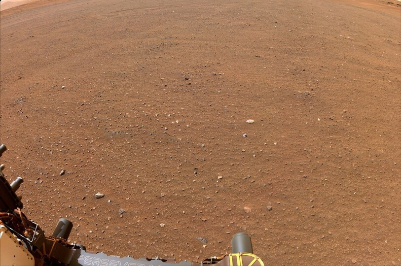 Flat, rocky terrain in Jezero Crater on Mars with edge of Perseverence Rover in lower left