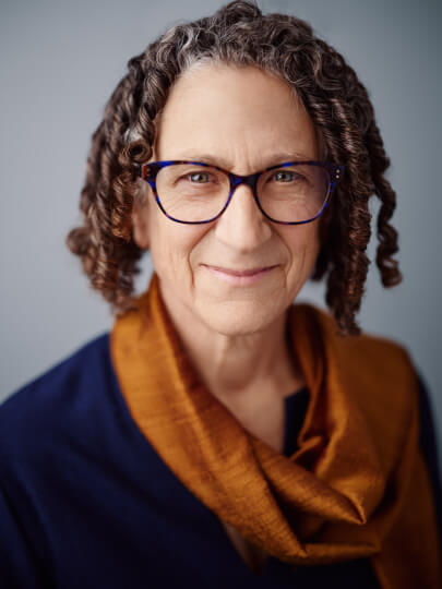 Smiling person with short curls and dark-framed glasses, wearing a navy top and an amber scarf, posed against a soft gray background.