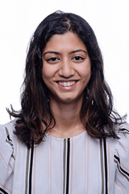 A person with shoulder-length brown hair, wearing a lavender blouse with black and pink stripes.