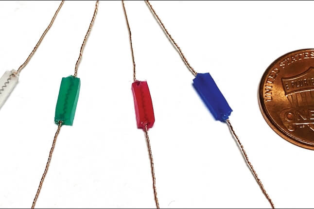 Four small hyrogel rectangles in clear, green, red, and blue are attached to four wires next to a penny, showing their miniscule size. 