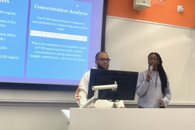 A male and a female Harvard student presenting behind a computer monitor, in front of a digital projection screen