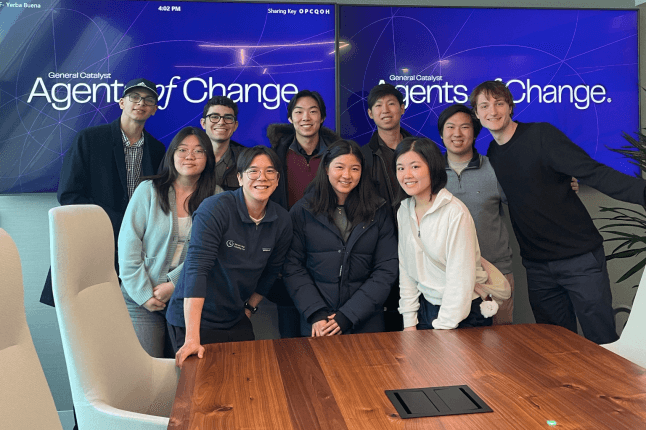 A group of Harvard SEAS students standing behind a wooden table, in front of a sign that says "Agents of Change"