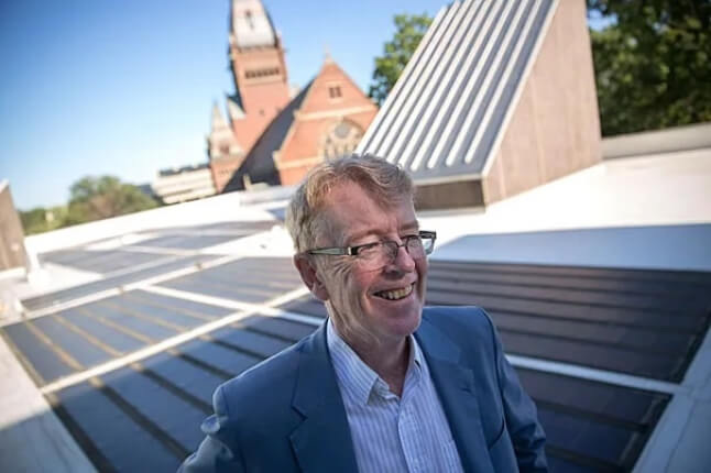 A man with glasses and light hair is smiling, dressed in a blue suit, and standing on a rooftop with solar panels. In the background, there is a building with a pointed clock tower and a blue sky.