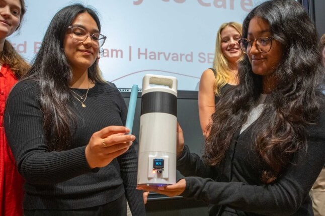 Two female Harvard SEAS students holding a grey container and blue medication vial