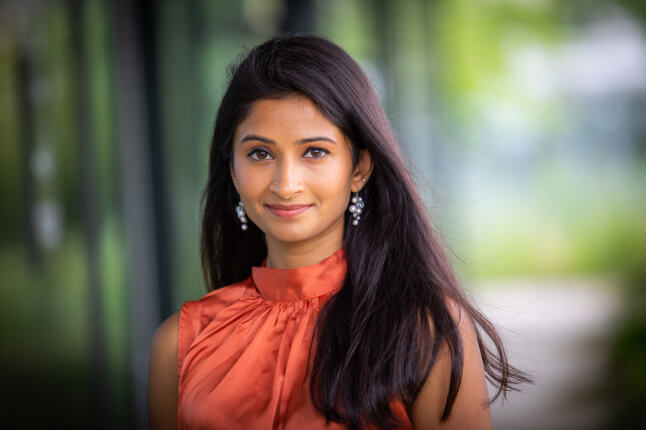 Woman with long dark hair wearing an orange blouse in an outdoor setting with a blurred green background