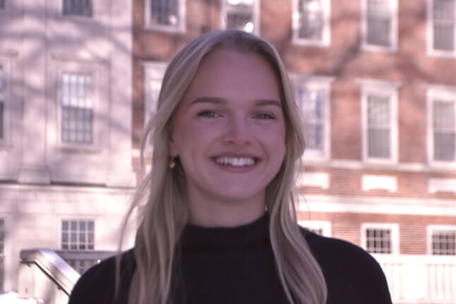 Portrait of a young woman with long blond hair wearing a dark sweater, in front of a red‑brick academic building.