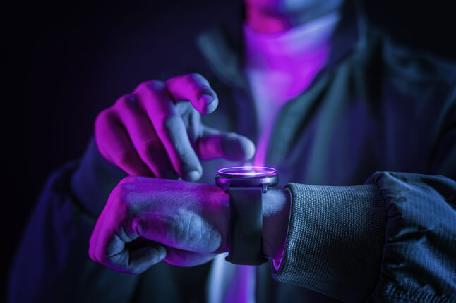 Close-up of a person’s hands in blue and purple lighting as they tap the screen of a smartwatch on their wrist.