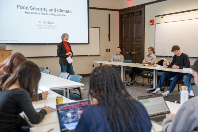  person stands at the front of a classroom giving a presentation titled “Food Security and Climate: Impossible Foods x GigaClimate” projected on a screen, while students sit around U‑shaped tables with laptops and notebooks, listening and taking notes.