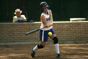 Woman in a white sleeveless jersey hits a yellow softball with a black baseball bat.