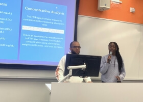 A male and a female Harvard student presenting behind a computer monitor, in front of a digital projection screen