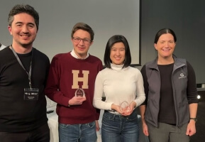 Four people standing in a line, one wearing a Harvard sweatshirt, two holding second-place statues