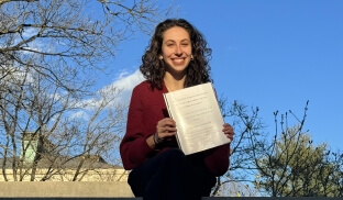 A female Harvard SEAS student wearing a red shirt and black pants sitting on a stone wall holding a laminated white booklet