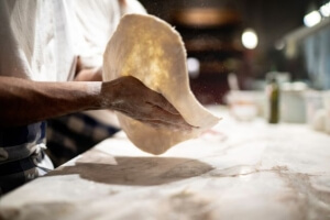 A person stretches pizza dough by hand over a marble countertop with flour visible on their hands.
