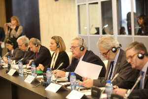 Group of suited delegates sit in a row at a conference table wearing headsets, reading documents, and speaking into microphones during a formal meeting