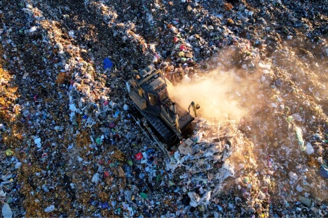 Aerial view of a bulldozer moving garbage at a landfill, kicking up dust among heaps of waste.